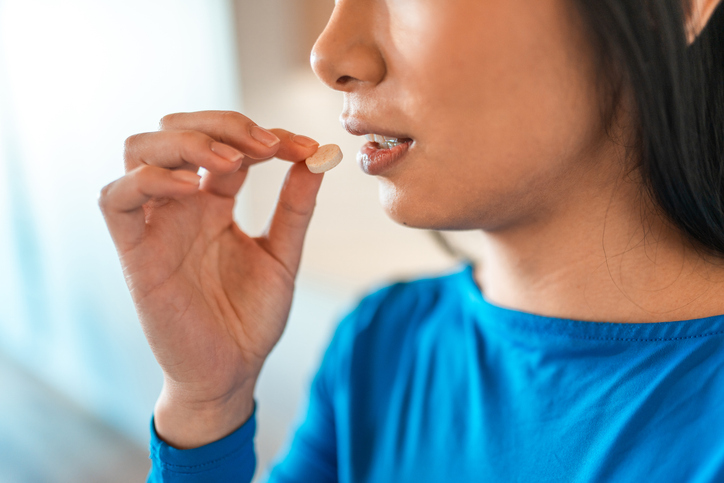 A young Asian woman holds a pill near her mouth, taking medication in a brightly lit indoor space.