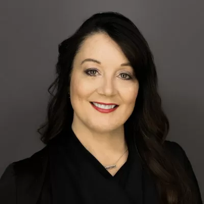 Portrait of a smiling woman with dark hair in a black blazer against a gray studio backdrop that conveys professionalism.