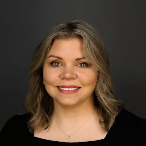 Head-and-shoulders portrait of a smiling woman with shoulder-length blonde hair in a black top against a dark backdrop.