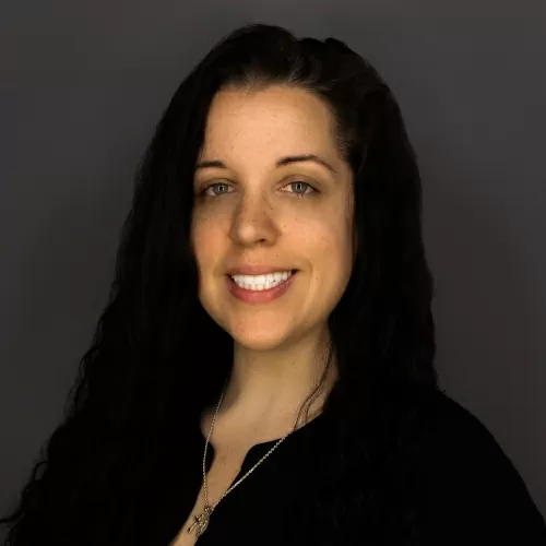 Smiling woman with long dark hair wearing a black top against a gray backdrop.