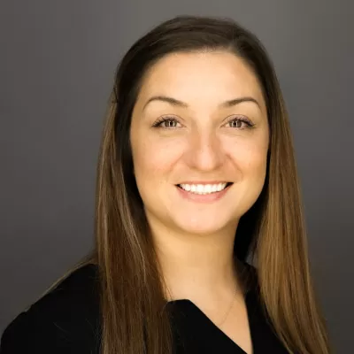 Professional headshot of a smiling woman with long brown hair, wearing a black top, against a neutral gray backdrop.
