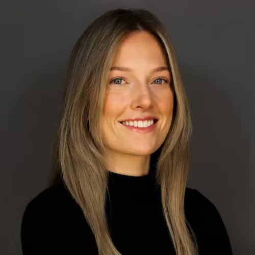 Smiling woman with long blonde hair in a black turtleneck, neutral gray background.