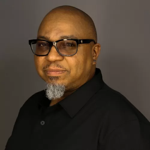 Professional headshot of a man wearing a black shirt and glasses against a gray studio background.