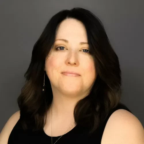Portrait of a woman with shoulder-length dark hair, wearing a black top, smiling softly against a gray studio background.