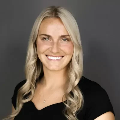 Professional headshot of a smiling blonde woman with wavy hair, wearing a black top, against a gray background.
