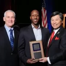 Three men in suits at an award ceremony; the center man holds a plaque while flanked by two others.