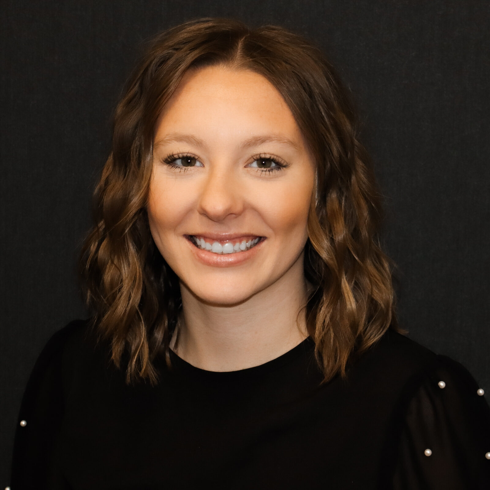 Portrait of a smiling woman with wavy brown hair, wearing a black top with pearl accents, against a dark background.