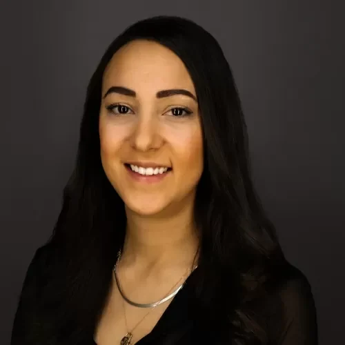 Portrait of a smiling woman with long dark hair and a silver necklace, against a dark gray background.