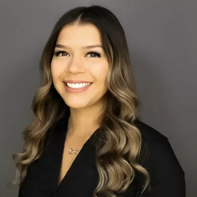 Head-and-shoulders portrait of a smiling woman with wavy, shoulder-length hair and a black blazer against a gray backdrop