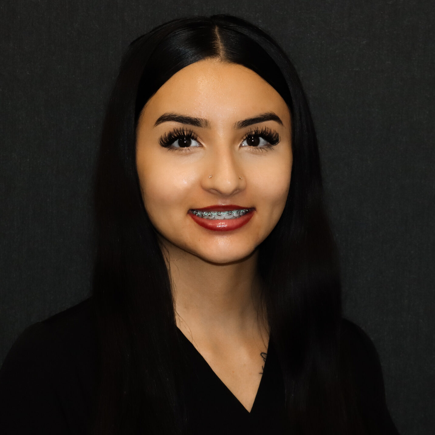 Portrait of a young woman with long black hair, wearing braces and nose studs, against a dark backdrop.