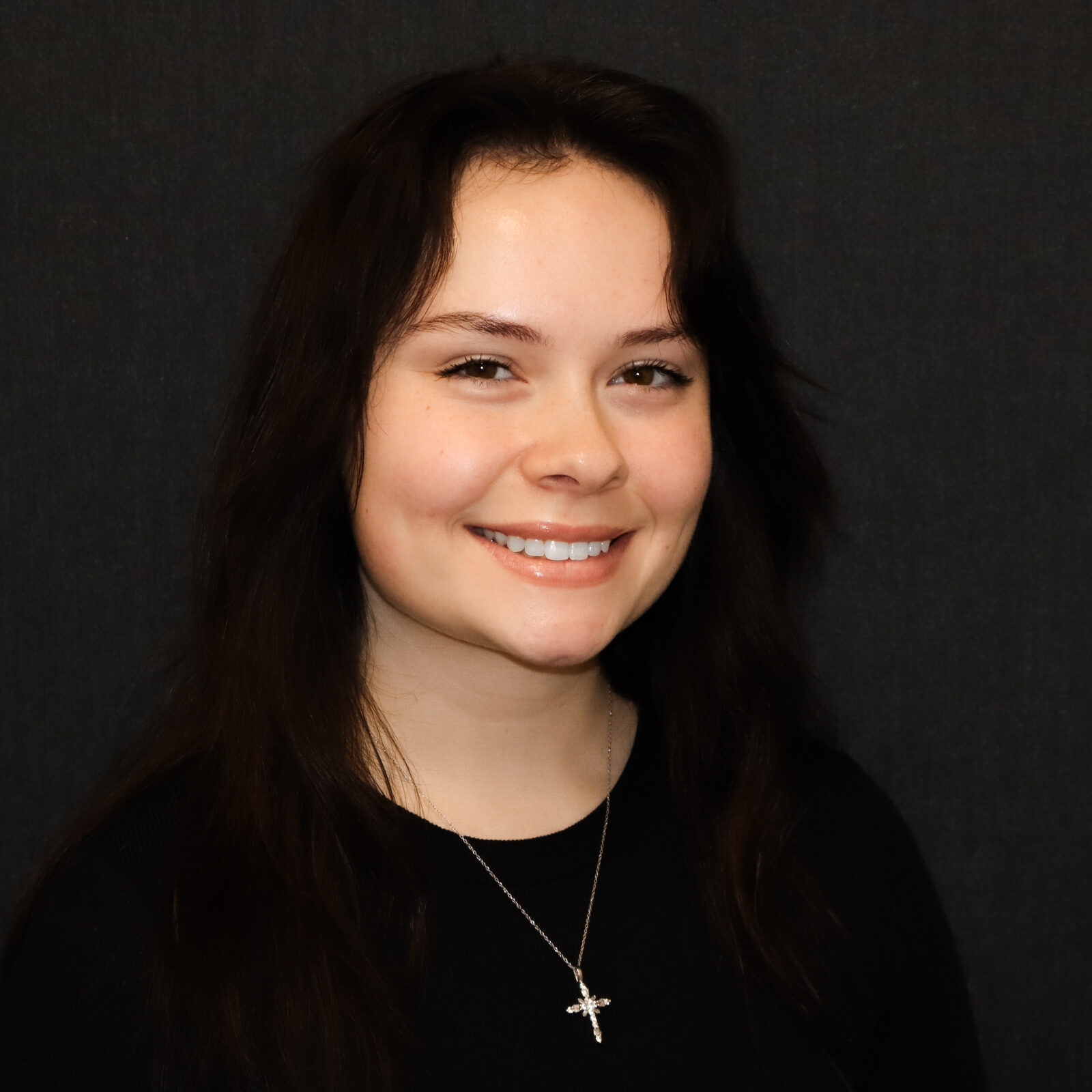 Young woman with long dark hair smiling against a dark backdrop, wearing a black top and a silver cross necklace.