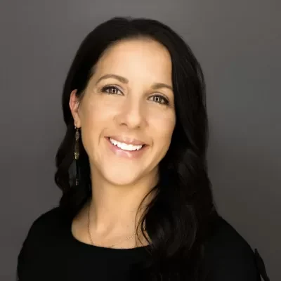 Smiling woman with dark hair, wearing a black top and drop earrings, against a gray background.